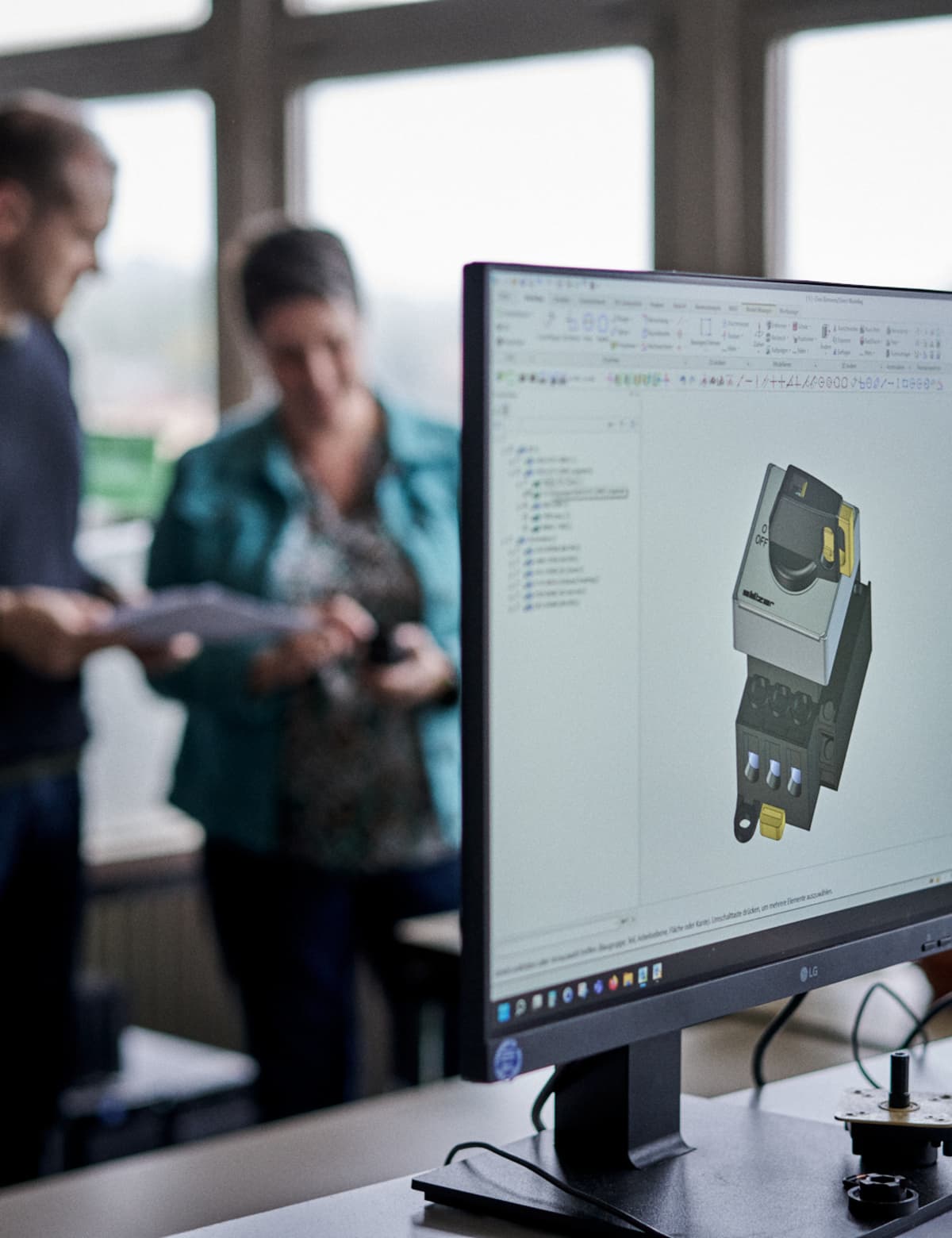 Zwei Mitarbeiter von Sälzer blettern in einem Katalog, im Vodergrund ist ein Bildschirm mit einer 3D-Ansicht von einem Nockenschalter